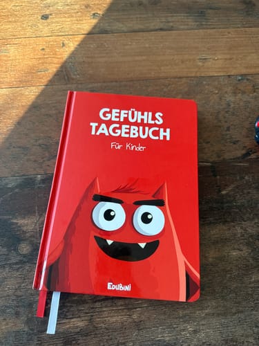 Rezensent zeigt das rote Gefühlstagebuch für Kinder mit einem freundlichen Monster auf dem Cover, das auf einem Holztisch liegt.