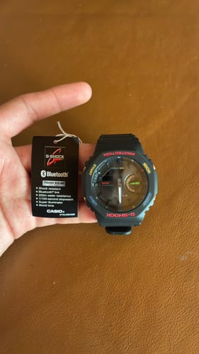 Reseñador sostiene en su mano el reloj Casio G-Shock negro con detalles en rojo, junto a la etiqueta del producto.