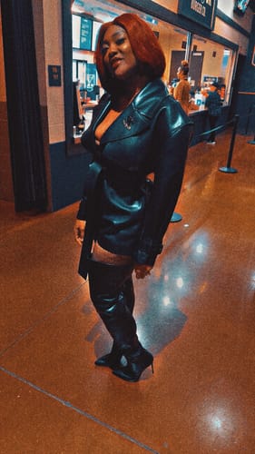 Reviewer standing indoors wearing the black Vegan Leather Trench Romper with matching thigh-high boots.