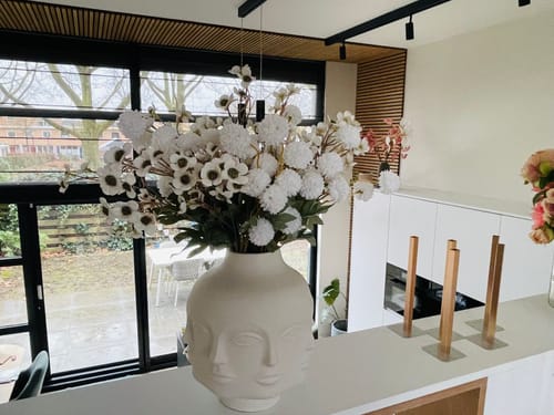 Recensent toont het Wax Arrangement - Wit in een decoratieve vaas in een modern interieur.