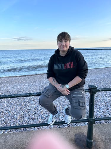 Reviewer sitting on a railing by a pebble beach, wearing the Dark Grey Cargo Pants V2 with large side pockets.