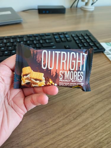 Customer holding a S'mores Peanut Butter Outright Protein Bar in their hand over a desk.