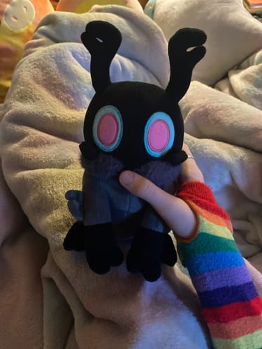 A reviewer's hand in a rainbow sleeve holds the soft Grey Scavenger plush against a fluffy blanket.