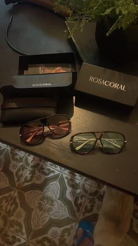 Cliente mostrando dois pares dos Óculos Veneza em cores diferentes, com a embalagem ao lado em uma mesa.