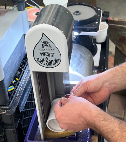 Customer using a silicon carbide sanding belt on a wet sander to shape a white cylindrical object.
