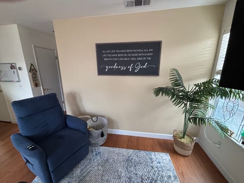 Customer's black Goodness Of God Faith Sign hanging in their living room, opposite a blue recliner.