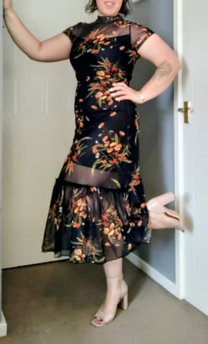 Customer wearing the black floral mesh midi dress, posing indoors with one leg lifted.