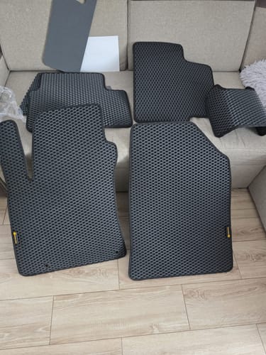 Recenzent prezentuje komplet czarnych dywaników samochodowych o strukturze plastra miodu, rozłożonych na podłodze i kanapie.