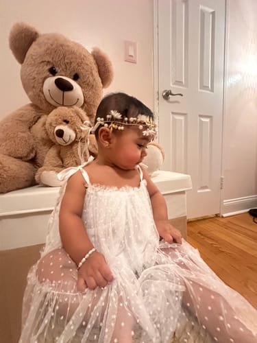 Reviewer's baby wearing the white Taya Tulle Dress, looking to the side while sitting in front of a teddy bear.