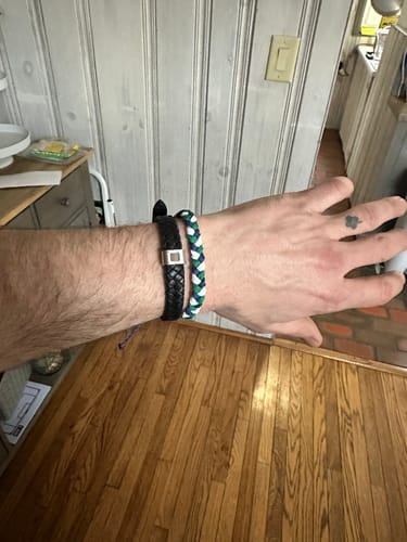 Reviewer's wrist wearing the woven blue, green, and white DAVID Bracelet next to a black bracelet.