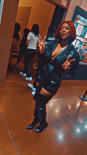 Reviewer posing in the black Vegan Leather Trench Romper and boots, making peace signs with both hands.
