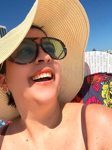 Cliente sorrindo em close-up, usando o Óculos Piemonte e um chapéu grande em um dia de sol.