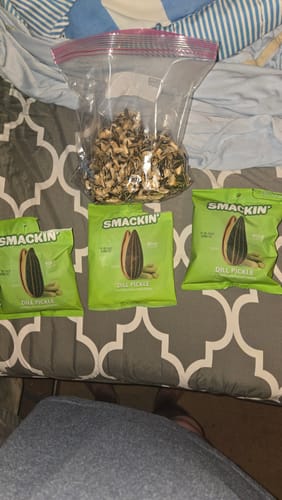 Reviewer's three bags of Dill Pickle sunflower seeds next to a large plastic bag of eaten shells on a bed.