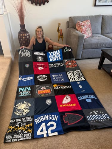 Reviewer proudly displaying her custom Premium T Shirt Quilt, made from various t-shirts, while sitting in her living room.
