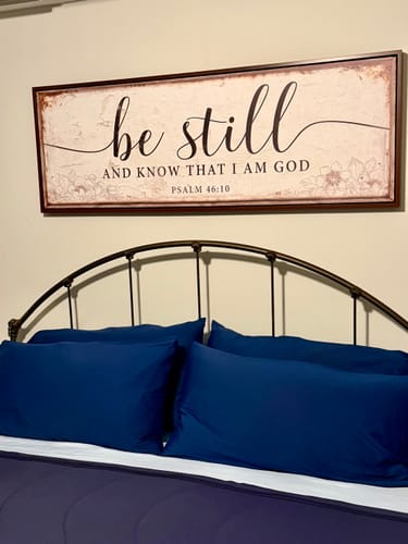 Customer's framed 'Be Still And Know That I Am God' canvas sign hanging on a wall above a bed.