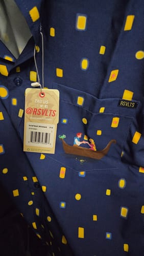 Reviewer's close-up of the navy Tangled "Lanterns" shirt, showing the pocket detail with characters in a boat and the RSVLTS tag.