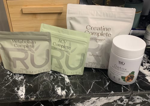 Reviewer's ACV Complete pouch shown with other TRU supplement bags and a container on a marble counter.