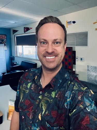Reviewer smiling while wearing the dark, dragon-patterned 'Scales 'n' Tails' short sleeve shirt indoors.