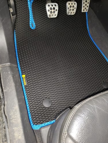 Recenzent pokazuje idealnie dopasowany czarny dywanik samochodowy z niebieskim obszyciem od strony kierowcy.