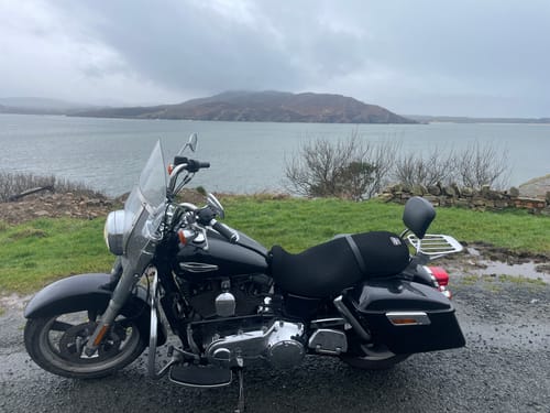 Customer's black Wind Rider Seat Cover installed on a motorcycle, parked with a scenic view of a lake and hills.