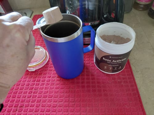Customer adds a scoop of Keto Activate powder to a beverage in a blue travel mug, with the container open next to it.
