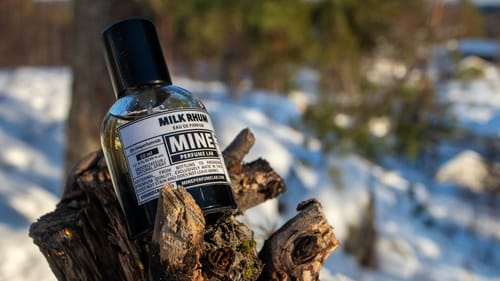 Profumo MILK RHUM del recensore, posato su un ceppo di legno in un paesaggio innevato.