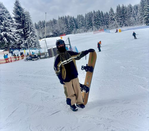Reviewer on a snowy slope wearing baggy multi-pocket snow pants while holding a snowboard.