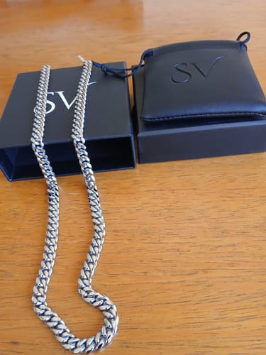 Cliente muestra la Cadena Cuban Link plateada en una mesa de madera junto a su caja y estuche negros.