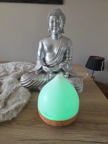 Klantfoto van de Unity 2.0 diffuser, verlicht met een groen licht, op een houten tafel voor een Boeddhabeeld.