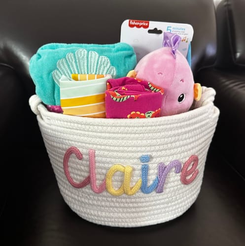 Customer's personalized white knitted basket with the name 'Claire' in multicolor letters, filled with baby gifts.