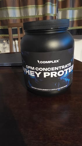 Reviewer's large black tub of Complex CFM Whey Protein sitting on a wooden table.