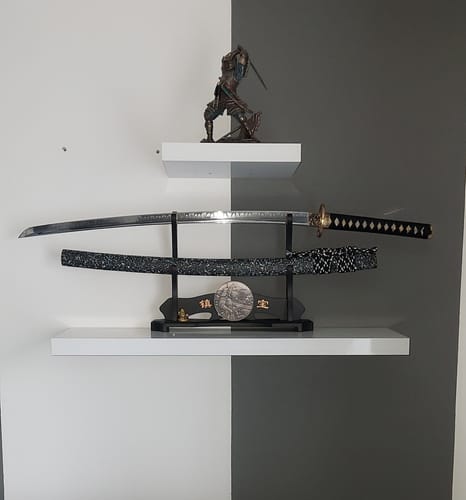 Reviewer's Star Stream Katana displayed on a stand, with the blade unsheathed above its patterned scabbard on a white shelf.