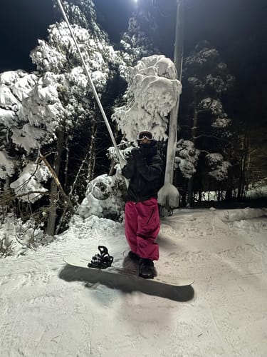 Reviewer in the oversized black snow jacket, standing on a snowboard on a snowy slope at night.