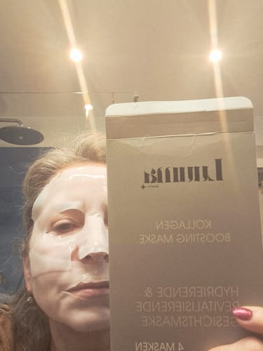 Kundin mit aufgetragener Kollagen Boosting Maske, die die Produktverpackung neben ihr Gesicht hält.