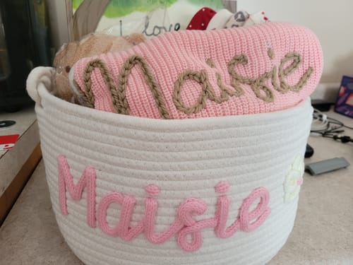 Customer's white knitted basket personalized with the name "Maisie" in pink yarn, filled with baby items and a matching pink blanket.