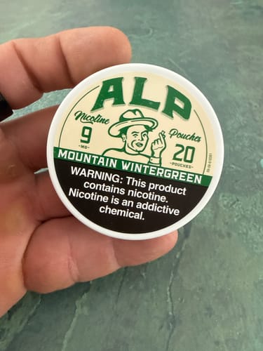 Customer holding a white can of ALP Mountain Wintergreen nicotine pouches against a green surface.
