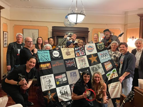 A group of customers holding up their large Premium T Shirt Quilt, made with various music-themed shirts, in a living room.