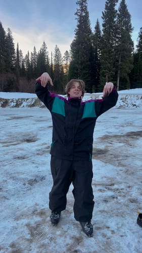 Reviewer in a snowy area pointing down at their baggy black snow pants to show the fit over their ski boots.