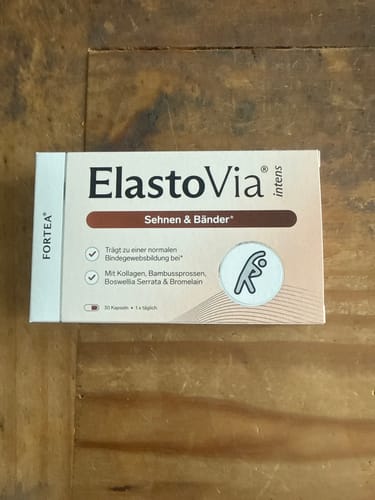Kunde zeigt die Produktverpackung von ElastoVia intens – Sehnen & Bänder auf einem Holztisch.