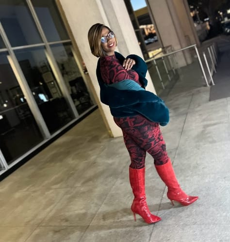 Customer posing outside at night in the snakeskin Naked Print Jumpsuit with a teal jacket and red boots.