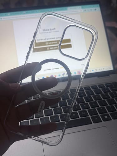 A customer's hand holding the clear Signature Magsafe Case for the iPhone 16 Pro Max in front of a laptop.