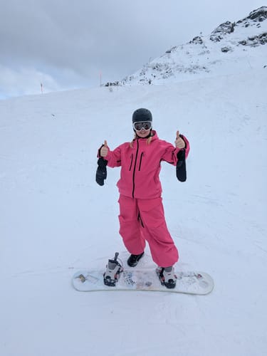 Reviewer gives a thumbs-up wearing the Rose Snow Jacket on a snowboard on a mountain.