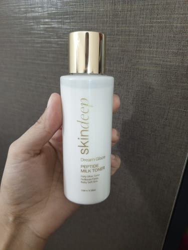 A reviewer's hand holding the white bottle of DREAM GLAZE Peptide Milk Toner against a dark background.