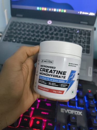 A customer holds a white tub of Wellcore Creatine Monohydrate in Watermelon Wave flavor over a keyboard.