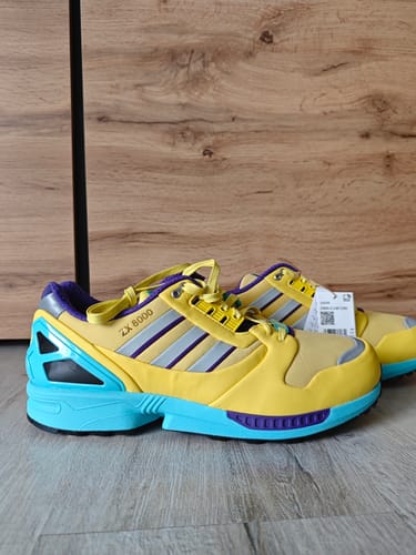Recensent toont een paar adidas ZX 8000 Consortium-sneakers in Yellow Blue Glow op een houten vloer.