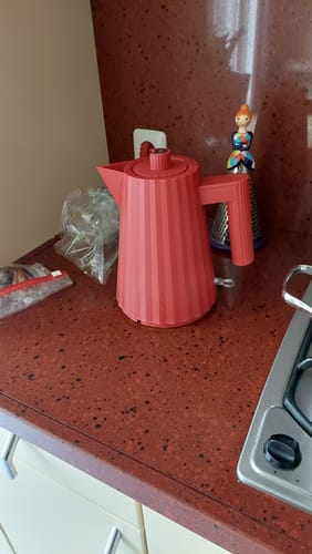 Beoordelaar toont de rode Alessi Plissé waterkoker op een donkerrood keukenblad.