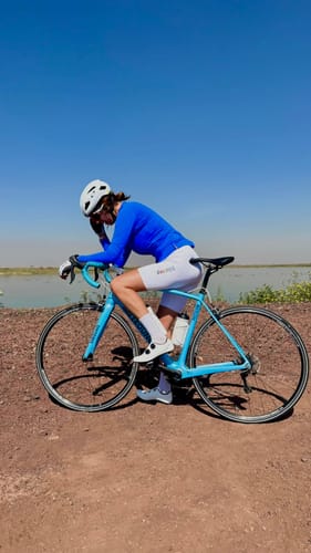Reviewer wearing the bright blue Vividness Jersey, leaning on their bicycle on a dirt path by the water.