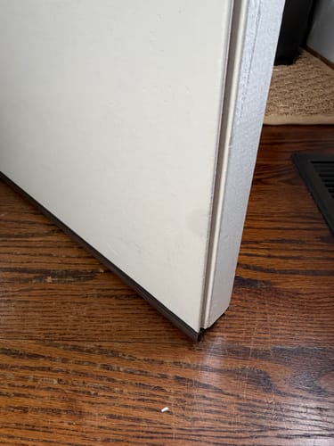 Close-up of a customer's installed corner seal at the bottom of a white door, sealing the gap above a dark wood floor.