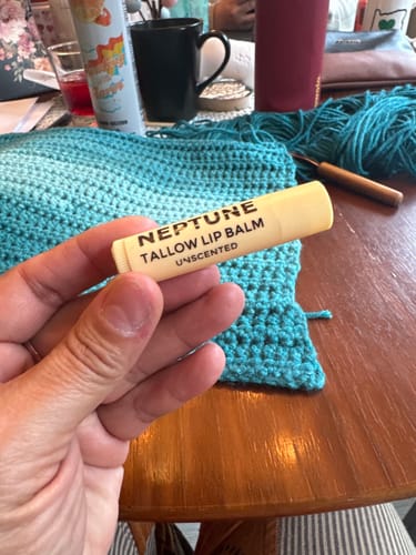 Reviewer's hand holding a tube of unscented Neptune Tallow Lip Balm over a wooden table with a crochet project.