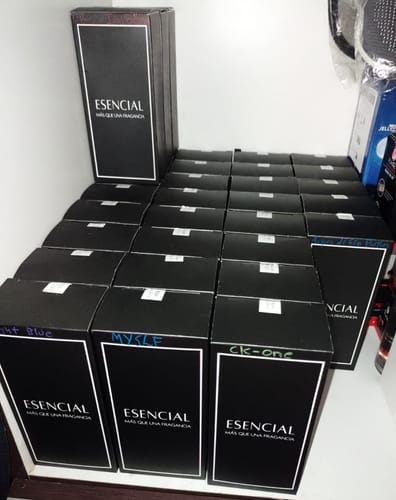 Cliente muestra su gran pedido de perfumes Esencial, con múltiples cajas negras apiladas en un estante.